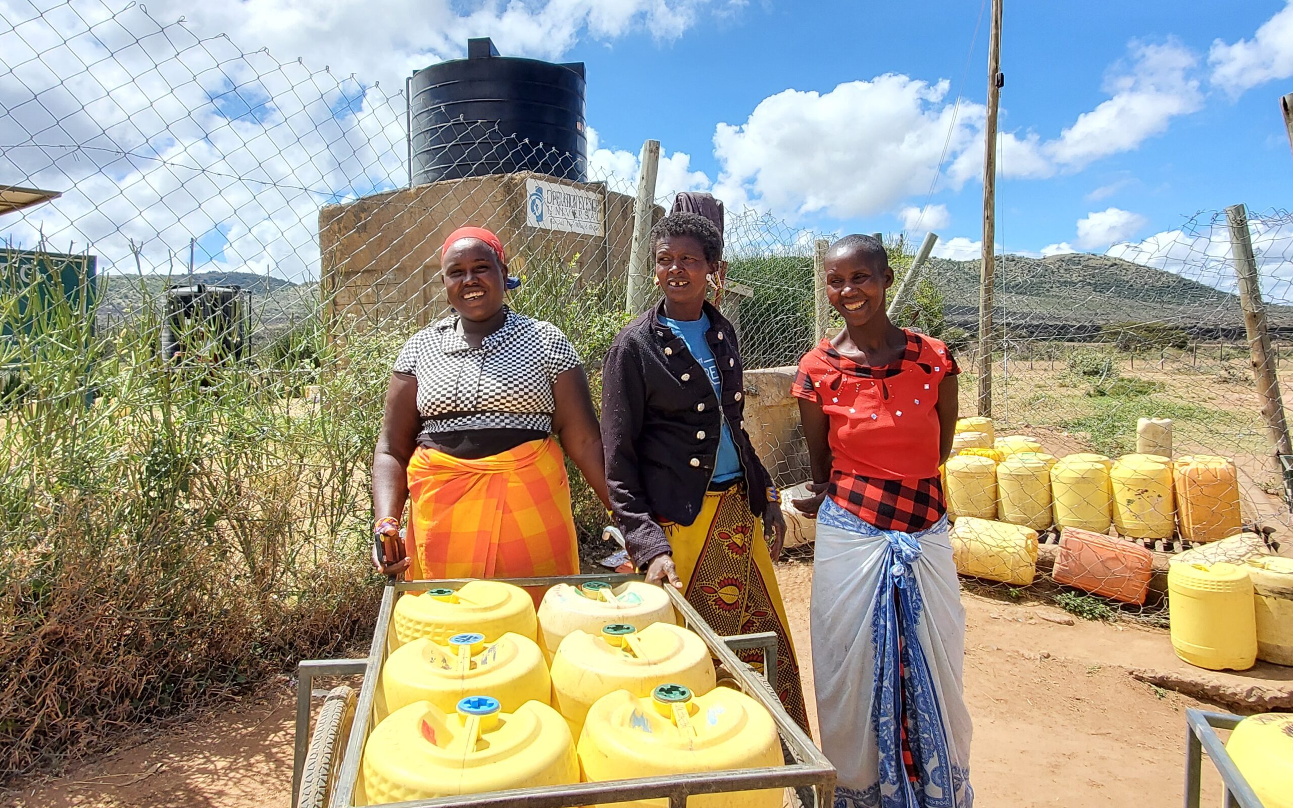 Three women smile as they collect water from a water tank