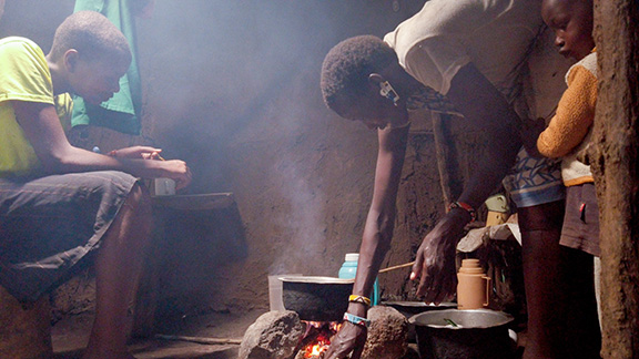 Woman bends over cooking fire with two children by her side.