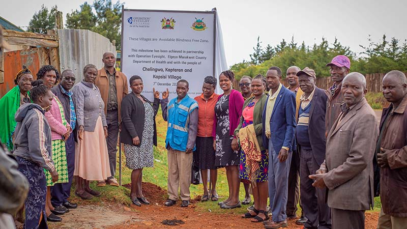 A group of people pose in front of a large poster announcing an Avoidable Blindness-Free Zone in a rural setting in Elgeyo Marakwet, Kenya.