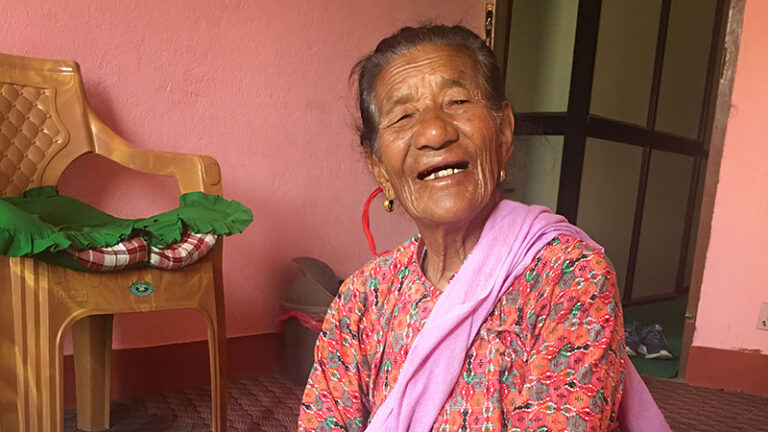 A senior woman smiles at the camera, seated on the floor of a house with pink walls.