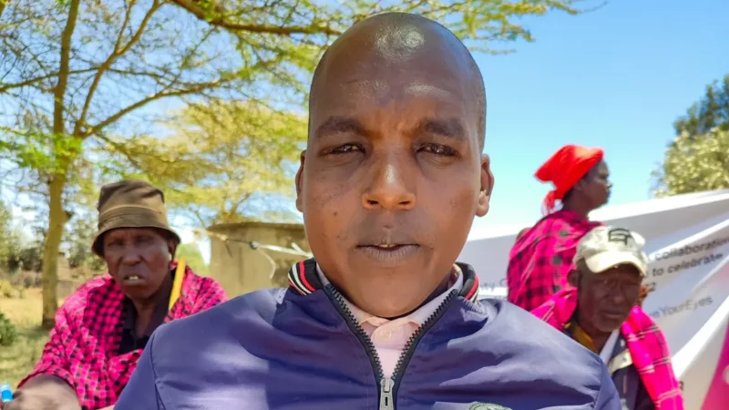 A young man wearing a blue jacket looks into the camera. There are people, trees and blue sky int the background.
