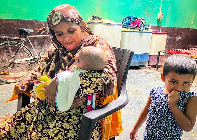A woman cradles an infant while sitting in a chair. A toddler stands next to them.