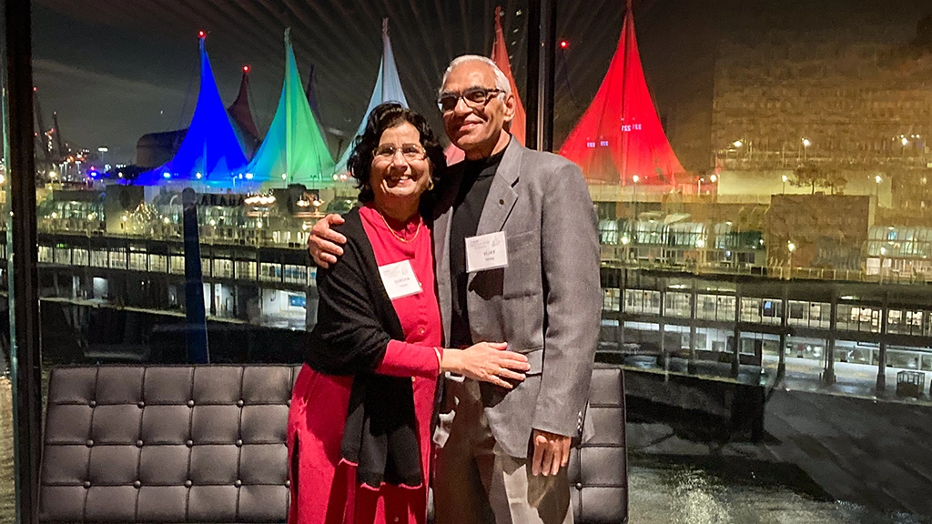 A man and woman pose with their arms around each other. In the background, you can see a building with colourful sails on top of it in the background.