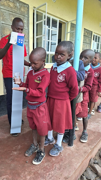 A group of uniformed schoolchildren stand in a queue. The boy at the front of the line holds a glass of water in one hand and a pill in the other.