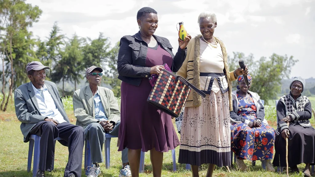 Two women stand outside with others sitting in the background. They are making a presentation and showing bead work.