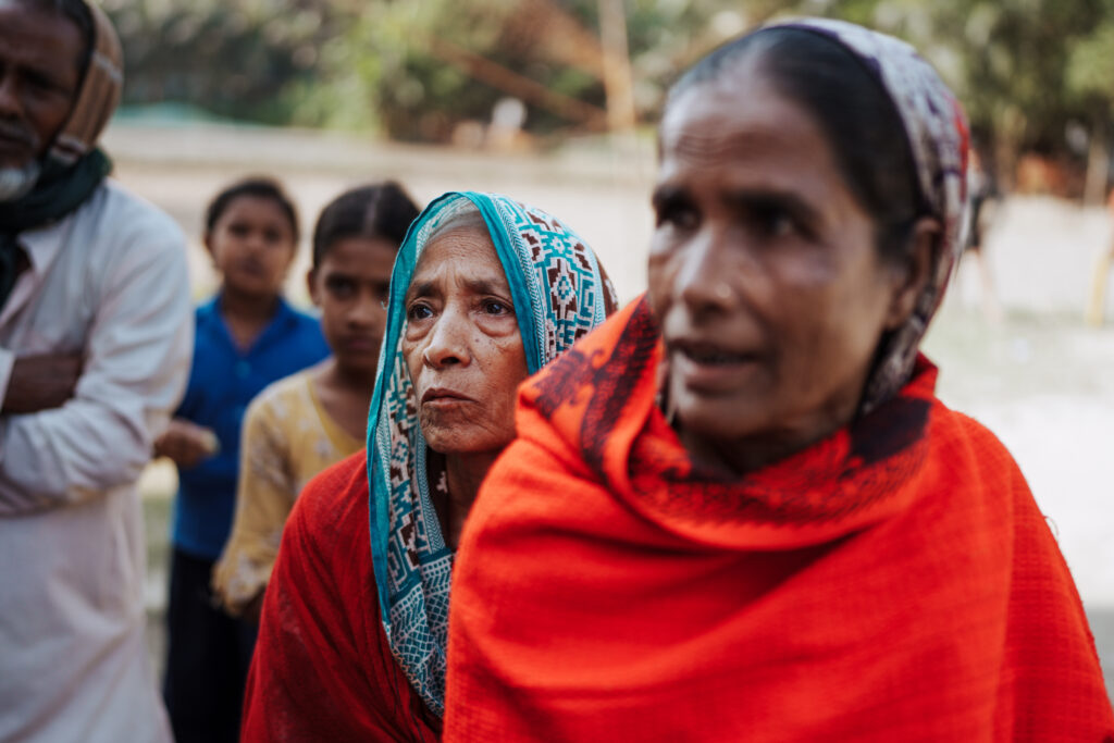 Gulbahar stands in line with other patients at the Operation Eyesight eye care camp in rural Bangladesh. She and another woman in the foreground wear red and orange shawls and patterned saris to cover their heads.