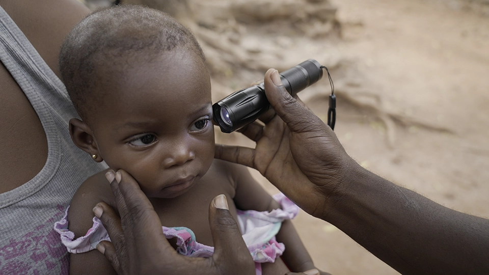 A baby sitting on her mother's knee has her eye examined with a flashlight. 