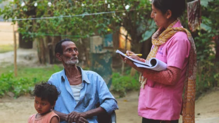 A woman ispeaks to a man sitting in a chair while taking notes in a notebook. A child leans against the man.